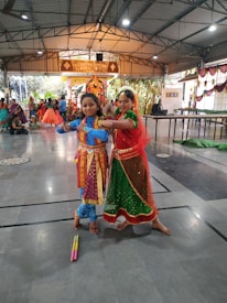Two people in traditional Indian attire are posed in the center of a large indoor hall. They are wearing colorful costumes, with intricate designs and traditional jewelry. Other people in similar attire can be seen in the background, and there are decorations hanging from the ceiling. The setting appears to be festive, possibly for a cultural or religious event.