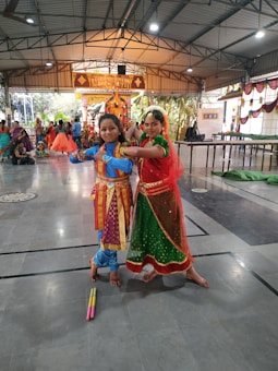 Two people in traditional Indian attire are posed in the center of a large indoor hall. They are wearing colorful costumes, with intricate designs and traditional jewelry. Other people in similar attire can be seen in the background, and there are decorations hanging from the ceiling. The setting appears to be festive, possibly for a cultural or religious event.