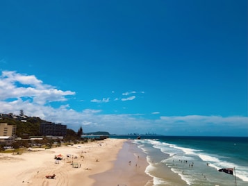 A sandy beach with people swimming and relaxing. The ocean stretches out to the horizon under a bright blue sky with scattered clouds. Buildings and greenery line the shore. An Australian flag is visible.