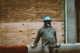 A person wearing a blue hard hat and a partially lowered face mask stands against a wooden construction barrier. The individual is dressed in a dark jacket and light gloves, embodying a scene of construction work or building maintenance.
