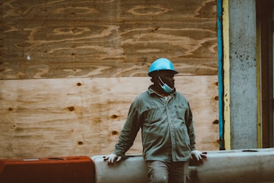 A person wearing a blue hard hat and a partially lowered face mask stands against a wooden construction barrier. The individual is dressed in a dark jacket and light gloves, embodying a scene of construction work or building maintenance.