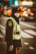 A traffic officer wearing a NYPD uniform stands on a city street. The officer is dressed warmly, wearing a thick hat and gloves, and a face mask. The background is blurred with traffic lights and cars indicating a busy urban environment.