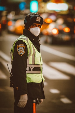 A traffic officer wearing a NYPD uniform stands on a city street. The officer is dressed warmly, wearing a thick hat and gloves, and a face mask. The background is blurred with traffic lights and cars indicating a busy urban environment.