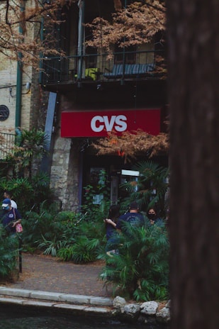 A CVS store entrance is seen surrounded by lush greenery, with trees and plants in the foreground. Several people are standing near the entrance, some wearing masks, indicating a casual urban environment. The setting feels serene due to the presence of greenery.