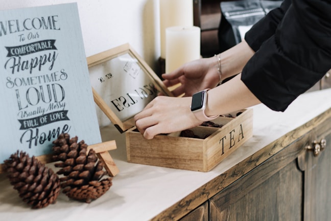 A person wearing a black sleeve and a smartwatch is opening a wooden box labeled 'TEA' on a countertop. Next to it is a decorative sign with words such as 'Welcome,' 'Comfortable,' and 'Happy.' Two pine cones are placed in front of the sign, and there are candles in the background.