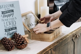 A person wearing a black sleeve and a smartwatch is opening a wooden box labeled 'TEA' on a countertop. Next to it is a decorative sign with words such as 'Welcome,' 'Comfortable,' and 'Happy.' Two pine cones are placed in front of the sign, and there are candles in the background.