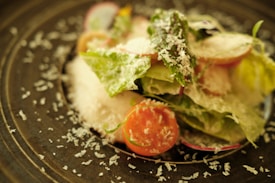 A fresh salad with crisp lettuce leaves, cherry tomatoes, radish slices, and a generous sprinkle of grated cheese is arranged on a dark plate. The focus on the salad emphasizes vibrant textures and freshness.