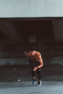 A muscular, shirtless man wearing black leggings and running shoes is captured in mid-stride, appearing to sprint forward. The setting is a dimly lit, urban environment with a concrete structure overhead, suggesting an underpass or bridge. The man's posture and focused expression convey determination and athleticism.