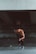 A muscular, shirtless man wearing black leggings and running shoes is captured in mid-stride, appearing to sprint forward. The setting is a dimly lit, urban environment with a concrete structure overhead, suggesting an underpass or bridge. The man's posture and focused expression convey determination and athleticism.