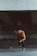 A muscular, shirtless man wearing black leggings and running shoes is captured in mid-stride, appearing to sprint forward. The setting is a dimly lit, urban environment with a concrete structure overhead, suggesting an underpass or bridge. The man's posture and focused expression convey determination and athleticism.