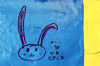 A simple drawing of a rabbit's face with long ears depicted on a blue background. The rabbit has a friendly expression. Next to it, the text 'Hi I (heart) MA SELF' is written in a casual, playful style. The image is bordered by a vertical yellow strip on the right.
