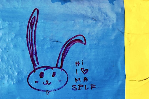 A simple drawing of a rabbit's face with long ears depicted on a blue background. The rabbit has a friendly expression. Next to it, the text 'Hi I (heart) MA SELF' is written in a casual, playful style. The image is bordered by a vertical yellow strip on the right.