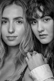 Two individuals are portrayed in close-up, black and white photography. One person has long, light hair while the other has dark hair with a fringe. They are positioned side by side, with the second person slightly behind. Their expressions are calm and neutral, and there are visible accessories such as rings.