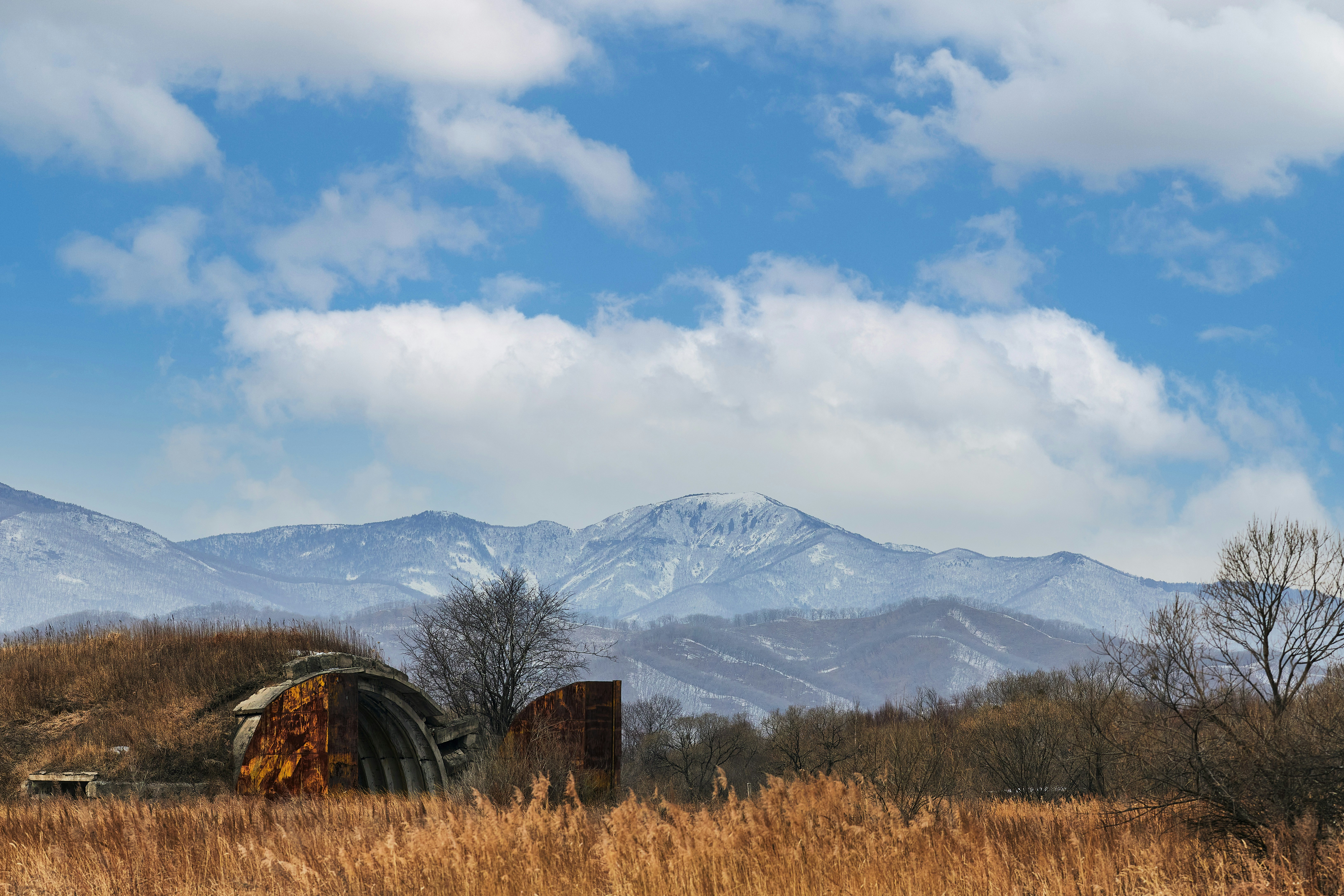 brown wooden house on brown grass field near snow covered mountain during daytime, Abandoned military hangar against the background of a field of yellow grass, snow-capped mountains, and white clouds