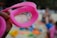 A pink swimming goggle is held up against a blurred beach background. The focus is on the goggles, which have water droplets on the lenses. The sandy beach and colorful toys or objects are visible in the background, slightly out of focus. The scene appears casual and relaxed, with a hand holding the goggles in the foreground.