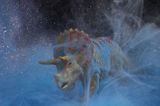 A model of a triceratops dinosaur is surrounded by swirling mist, creating an ethereal and mysterious ambiance. The figure is detailed with a textured skin painted in shades of blue and gold, with hints of red, and it seems to be emerging from a dreamy, cosmic-like environment.