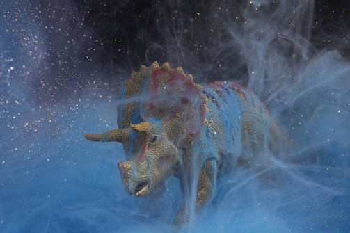 A model of a triceratops dinosaur is surrounded by swirling mist, creating an ethereal and mysterious ambiance. The figure is detailed with a textured skin painted in shades of blue and gold, with hints of red, and it seems to be emerging from a dreamy, cosmic-like environment.