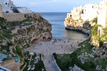 A picturesque cove flanked by rugged cliffs leads to a calm, blue sea. The sandy beach is scattered with people enjoying leisure activities. The cliffs are dotted with greenery, and there are buildings on the clifftop, some of which appear to be a hotel. Various beach furniture like umbrellas and chairs can be seen arranged below the cliff.