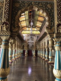 An ornate hall with intricate arches and decorative columns, featuring dominant gold and teal tones. The ceiling showcases stained glass panels, allowing a warm light to filter through, while a grand chandelier hangs centrally. The floor has a polished reflective surface, enhancing the room's opulent atmosphere.