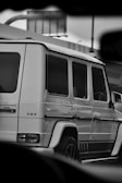 A luxurious SUV, specifically a Mercedes-Benz G-Class, is parked with a partial view of its side profile. The image is in black and white, highlighting the vehicle's contours and features. The background includes urban elements like street signs and part of another vehicle.