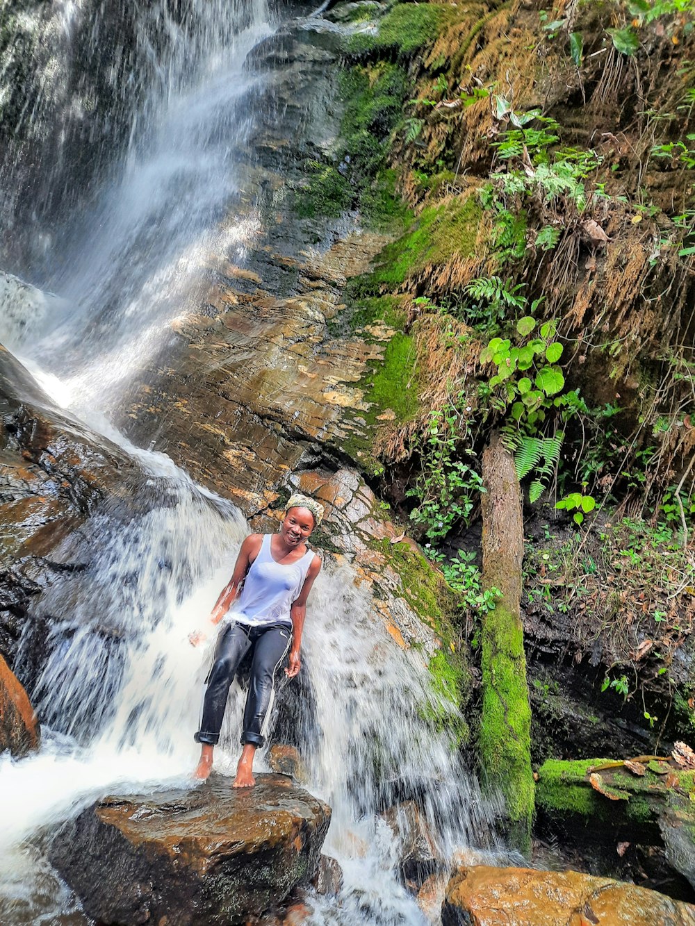 woman in white shirt and blue denim jeans standing on rock near waterfalls during daytime