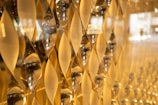 A close-up view of an artistic wall installation featuring an array of geometric, hourglass-shaped glass elements. These elements capture and reflect light, creating a golden, shimmering effect across the surface.