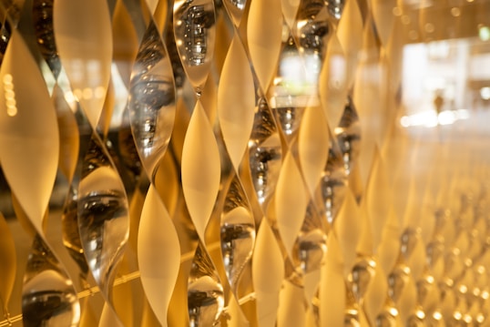 A close-up view of an artistic wall installation featuring an array of geometric, hourglass-shaped glass elements. These elements capture and reflect light, creating a golden, shimmering effect across the surface.