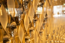 A close-up view of an artistic wall installation featuring an array of geometric, hourglass-shaped glass elements. These elements capture and reflect light, creating a golden, shimmering effect across the surface.