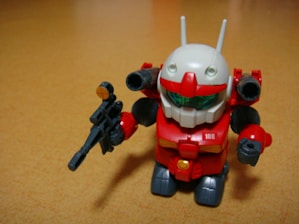 A small toy robot figure with a vibrant red body, white head, and green visor stands on a wooden surface. The robot holds a black weapon in its left hand and has antenna-like protrusions on its head.