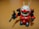 A small toy robot figure with a vibrant red body, white head, and green visor stands on a wooden surface. The robot holds a black weapon in its left hand and has antenna-like protrusions on its head.