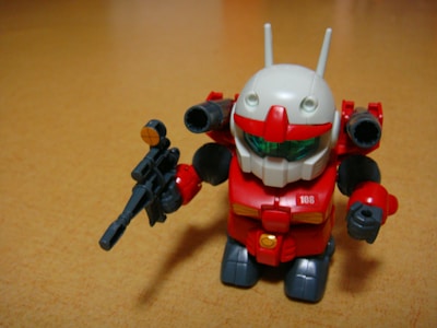 A small toy robot figure with a vibrant red body, white head, and green visor stands on a wooden surface. The robot holds a black weapon in its left hand and has antenna-like protrusions on its head.