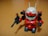 A small toy robot figure with a vibrant red body, white head, and green visor stands on a wooden surface. The robot holds a black weapon in its left hand and has antenna-like protrusions on its head.