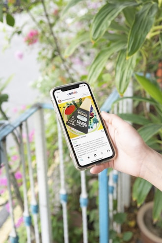 A hand holds a smartphone displaying a social media post of a vegan cheese product called 'Violife Wedge'. The phone screen shows a colorful image of the product with text below providing details. In the background, there are green plants and a railing with blue accents.