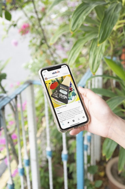 A hand holds a smartphone displaying a social media post of a vegan cheese product called 'Violife Wedge'. The phone screen shows a colorful image of the product with text below providing details. In the background, there are green plants and a railing with blue accents.