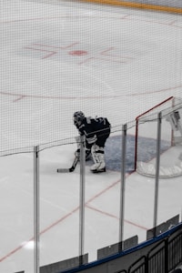 A hockey goalie dressed in dark gear is positioned in front of the net, standing on ice inside an arena. The goal is surrounded by protective glass panels and the surface of the ice is marked with red and blue lines. The image is taken from a distance, with the goalie appearing ready for action.