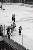 Several ice hockey players in blue jerseys are seen on the ice rink, with some standing near the boards and others closer to the center. Three referees or officials in black and white striped uniforms are standing together talking. In the background, a person is cleaning the ice with a shovel.