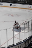 A hockey goalie wearing a red and white uniform is kneeling on the ice in front of a net. The rink is bordered by clear glass panels, and there are advertisements on the boards. The player has their back turned, with 'Johansson' and the number '31' visible on the jersey.