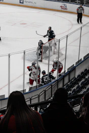 A hockey match takes place on an ice rink with players from two teams wearing different uniforms. The game appears intense as players are engaged near a goal net. Spectators watch from the stands while a referee observes the game closely.