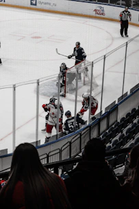 A hockey match takes place on an ice rink with players from two teams wearing different uniforms. The game appears intense as players are engaged near a goal net. Spectators watch from the stands while a referee observes the game closely.