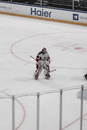 A hockey player, dressed in full gear, stands on the ice near the goal. They are holding a hockey stick and appear to be a goalie based on their gear and positioning. The rink has advertisements around its perimeter, including a visible 'Haier' sign.