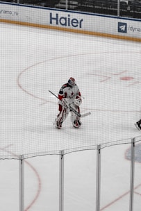 A hockey player, dressed in full gear, stands on the ice near the goal. They are holding a hockey stick and appear to be a goalie based on their gear and positioning. The rink has advertisements around its perimeter, including a visible 'Haier' sign.