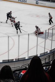 A hockey game is in progress with players in action near the goal. Two players in black jerseys are seen moving towards the goal, where an opposing player in a red and white jersey is positioned as the goalie. Two referees are also on the ice, watching the game closely. The scene is viewed from behind a spectator in the stands, with the ice surrounded by protective glass.