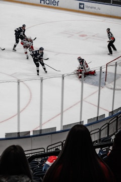 A hockey game is in progress with players in action near the goal. Two players in black jerseys are seen moving towards the goal, where an opposing player in a red and white jersey is positioned as the goalie. Two referees are also on the ice, watching the game closely. The scene is viewed from behind a spectator in the stands, with the ice surrounded by protective glass.