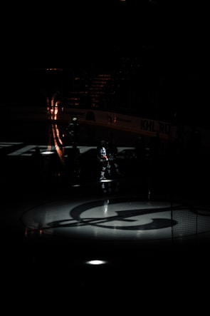 A dimly lit ice hockey rink with an illuminated logo on the ice. Shadows and light patterns create a dramatic atmosphere. The silhouettes of players are barely visible, standing around the rink.