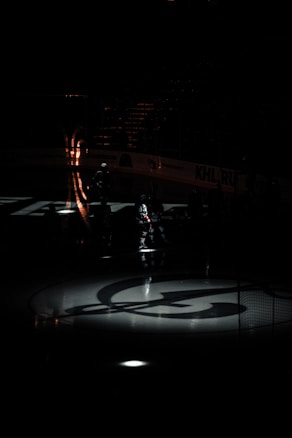 A dimly lit ice hockey rink with an illuminated logo on the ice. Shadows and light patterns create a dramatic atmosphere. The silhouettes of players are barely visible, standing around the rink.