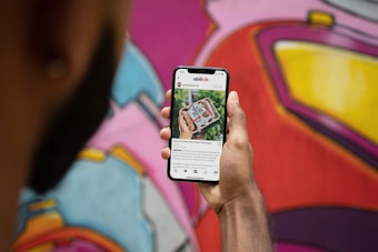 A person holds a smartphone displaying a social media app, with a focus on an image of packaged plant-based sausages. The background features a colorful mural with abstract geometric shapes and vibrant colors.