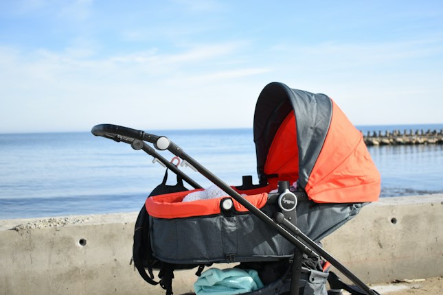 A baby stroller with a bright orange canopy is parked beside a concrete path overlooking a calm, blue ocean. The sky is clear with light clouds, and a breakwater is visible in the distance.