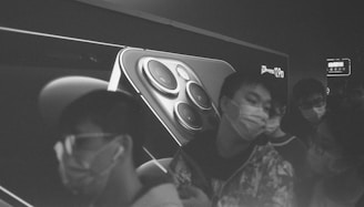 A black and white image features a large advertisement of an iPhone 12 Pro on a billboard, with a close-up of the phone's camera lenses. In the foreground, several people wearing masks are captured in motion, adding a sense of urgency and movement.