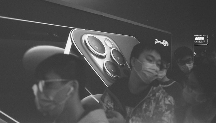 A black and white image features a large advertisement of an iPhone 12 Pro on a billboard, with a close-up of the phone's camera lenses. In the foreground, several people wearing masks are captured in motion, adding a sense of urgency and movement.