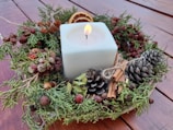A square white candle is surrounded by a festive wreath made of pinecones, cinnamon sticks, acorns, dried berries, and slices of dried orange. The arrangement sits on a wooden surface.